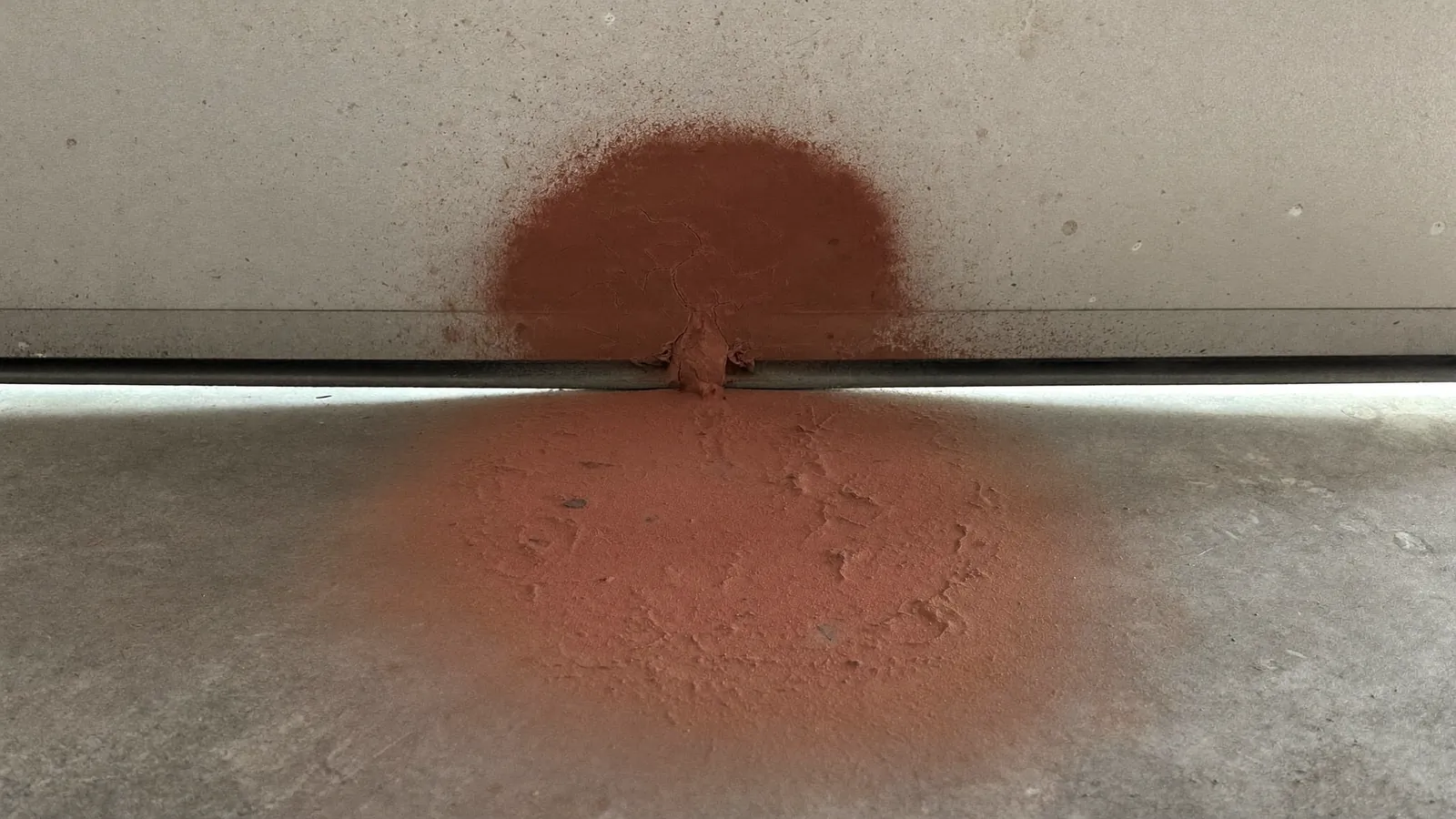 Fan of fine red-brown dust blown under a garage door with a worn bottom seal, spread across the gray concrete floor inside the garage
