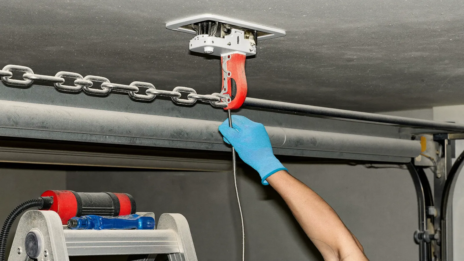 Technician's hands working on a ceiling-mounted chain-drive garage door opener, the rail and housing filmed with fine pale dust, a drill and new gear kit on a ladder tray