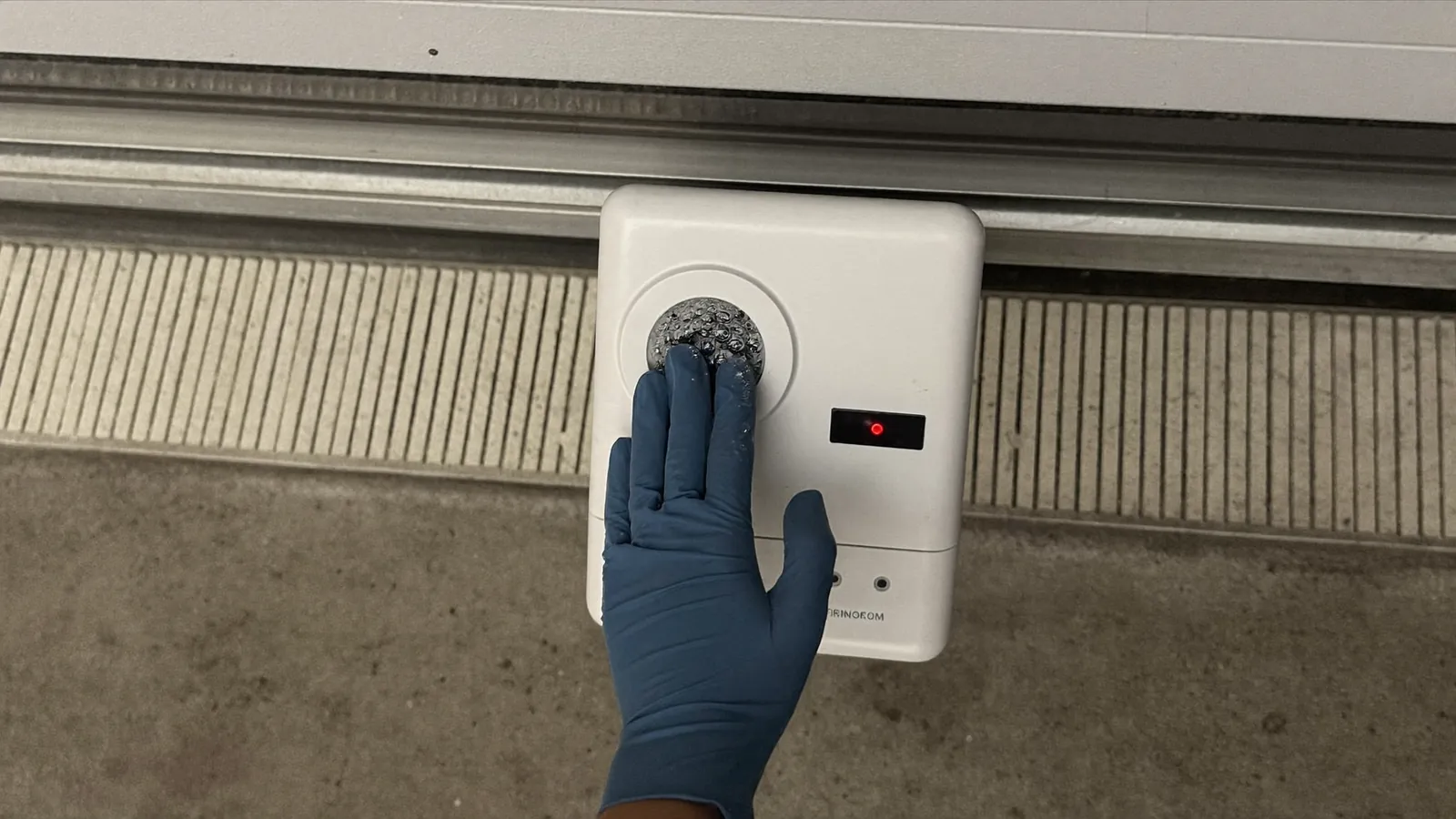 Gloved hand holding a garage door photo-eye safety sensor against the track near the floor, its lens dulled by a film of dust, indicator light showing