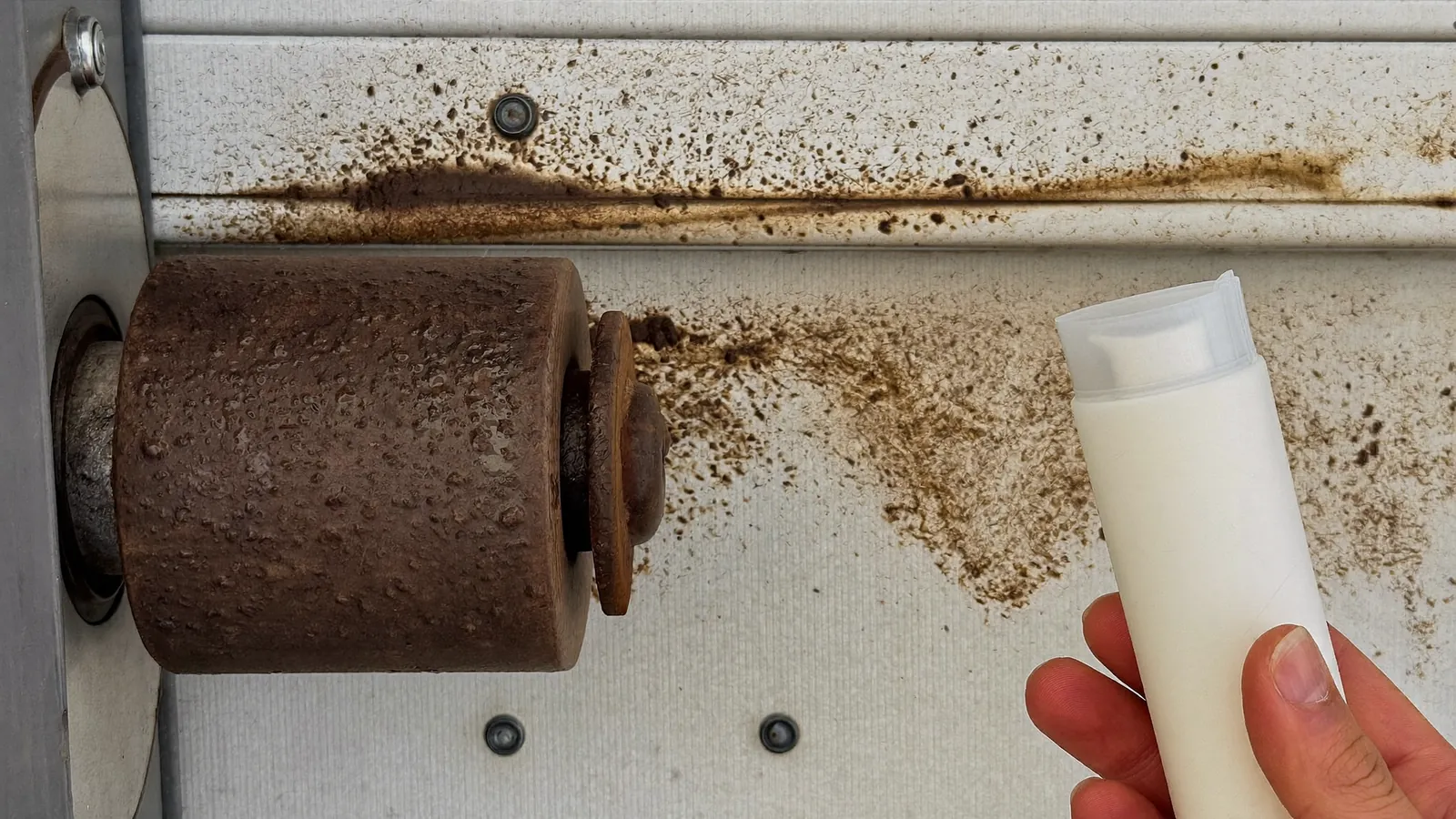Close-up of a garage door roller in a vertical track caked with a dark paste of grease and wind-blown grit, a clean replacement nylon roller held beside it