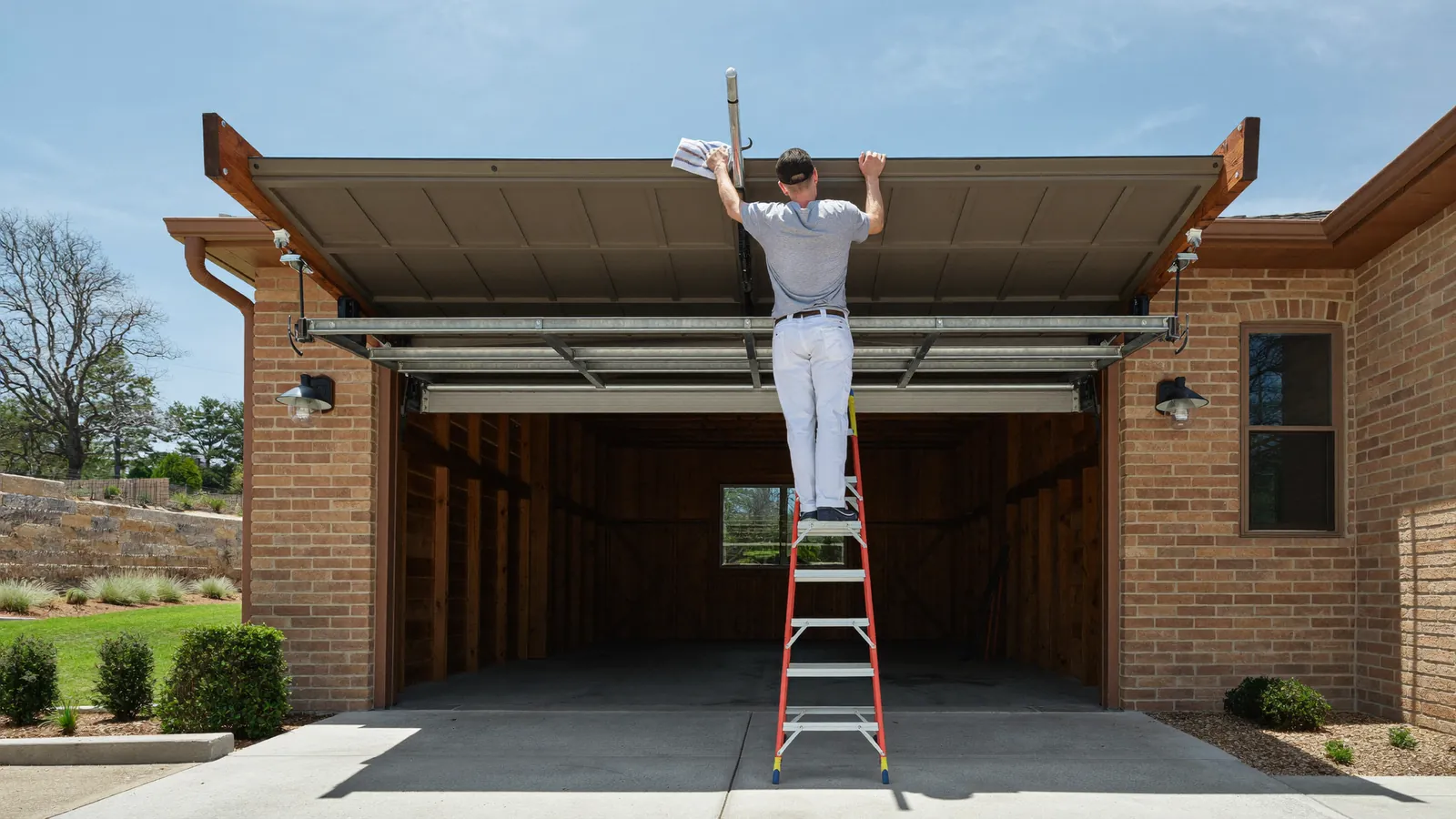 Garage door technician on a step ladder cleaning grit out of the roller track above an open double garage door of a tan brick ranch home in Lubbock, Texas, flat West Texas sky behind