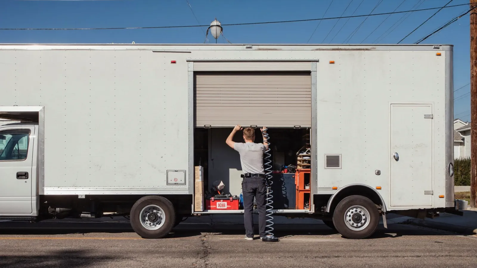Garage door technician reaching into the open side of a plain white service truck for parts, parked on a residential street in Lubbock, Texas, under a flat open sky