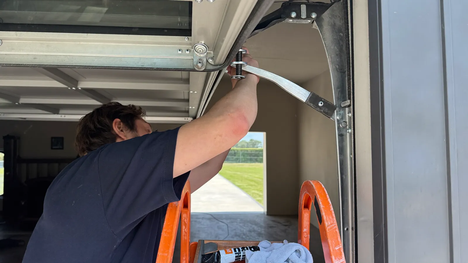 Technician on a ladder tightening a hinge bolt on a sectional garage door, rollers freshly replaced, a small container of lubricant and a rag on the ladder tray