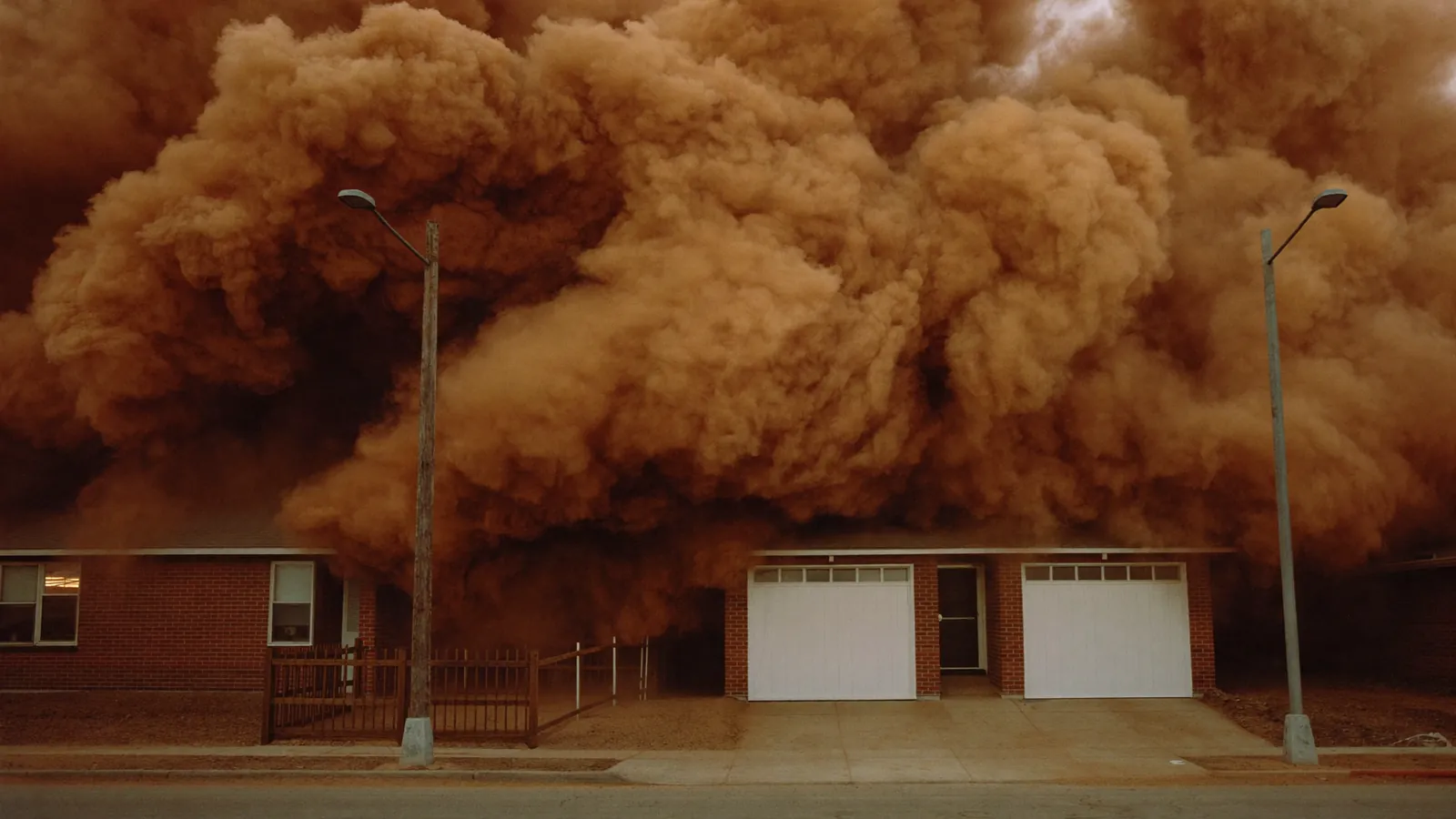 Towering brown wall of wind-blown dust rolling toward a Lubbock neighborhood of one-story brick homes with garage doors facing the street, streetlights coming on