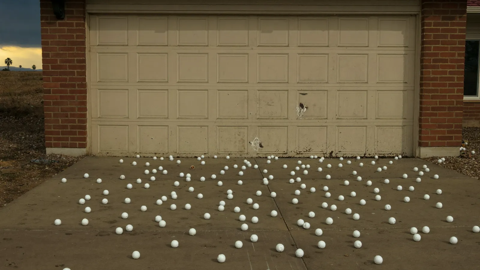 Golf-ball-size hailstones scattered across a wet concrete driveway in front of a dented white garage door after a West Texas storm, sun breaking through